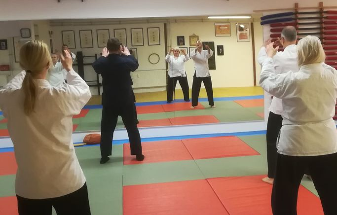 Gruppe von Personen in weißen Anzügen übt Bewegungstraining in einem Übungsraum.
